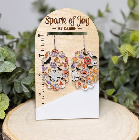 A pair of Halloween-themed dangle earrings displayed on a wooden card labeled 'Spark of Joy BY CARRIE'. The earrings feature a collage of small colorful icons including ghosts, pumpkins, candy corn, and peace signs.