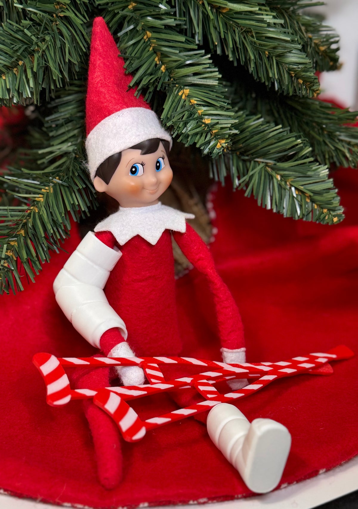 Elf doll wearing a white 3D printed arm cast and leg cast, standing with red crutches under a Christmas tree