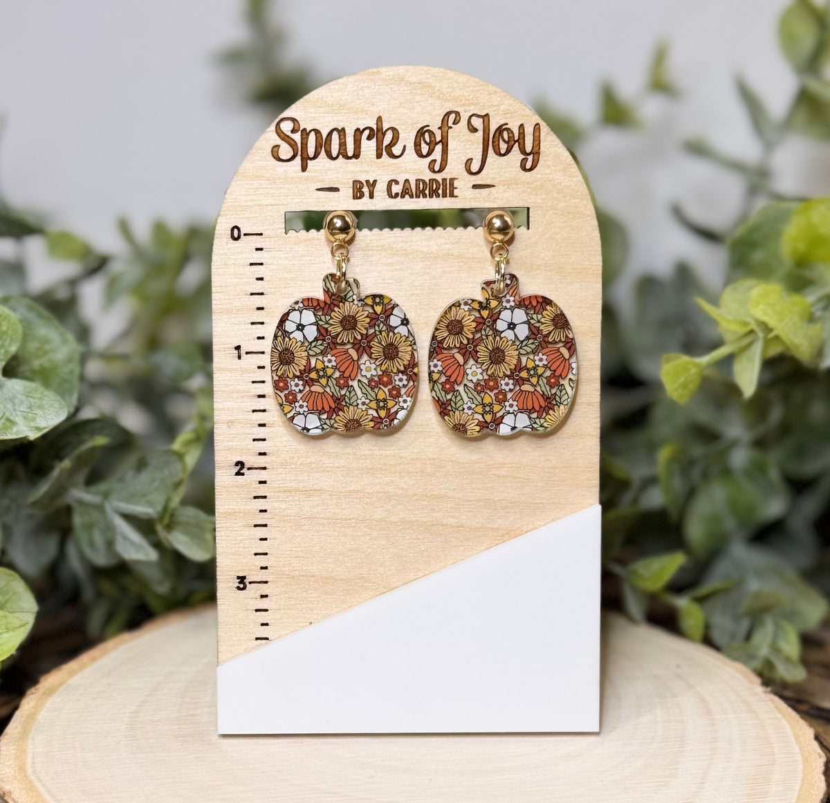 A pair of pumpkin-shaped dangling earrings with a colorful floral pattern, displayed on a wooden stand labeled 'Spark of Joy by Carrie' with a size ruler, set against a blurred green foliage background.