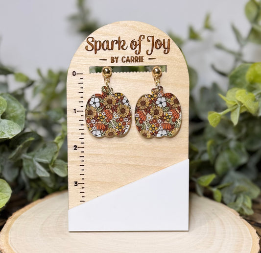 A pair of pumpkin-shaped dangling earrings with a colorful floral pattern, displayed on a wooden stand labeled 'Spark of Joy by Carrie' with a size ruler, set against a blurred green foliage background.