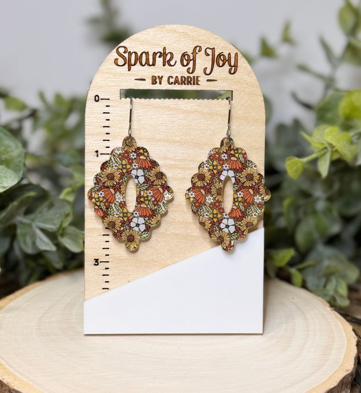 A pair of ornate dangling earrings displayed on a wooden stand labeled 'Spark of Joy BY CARRIE.' The earrings have a scalloped floral pattern in orange, brown, white, and green.