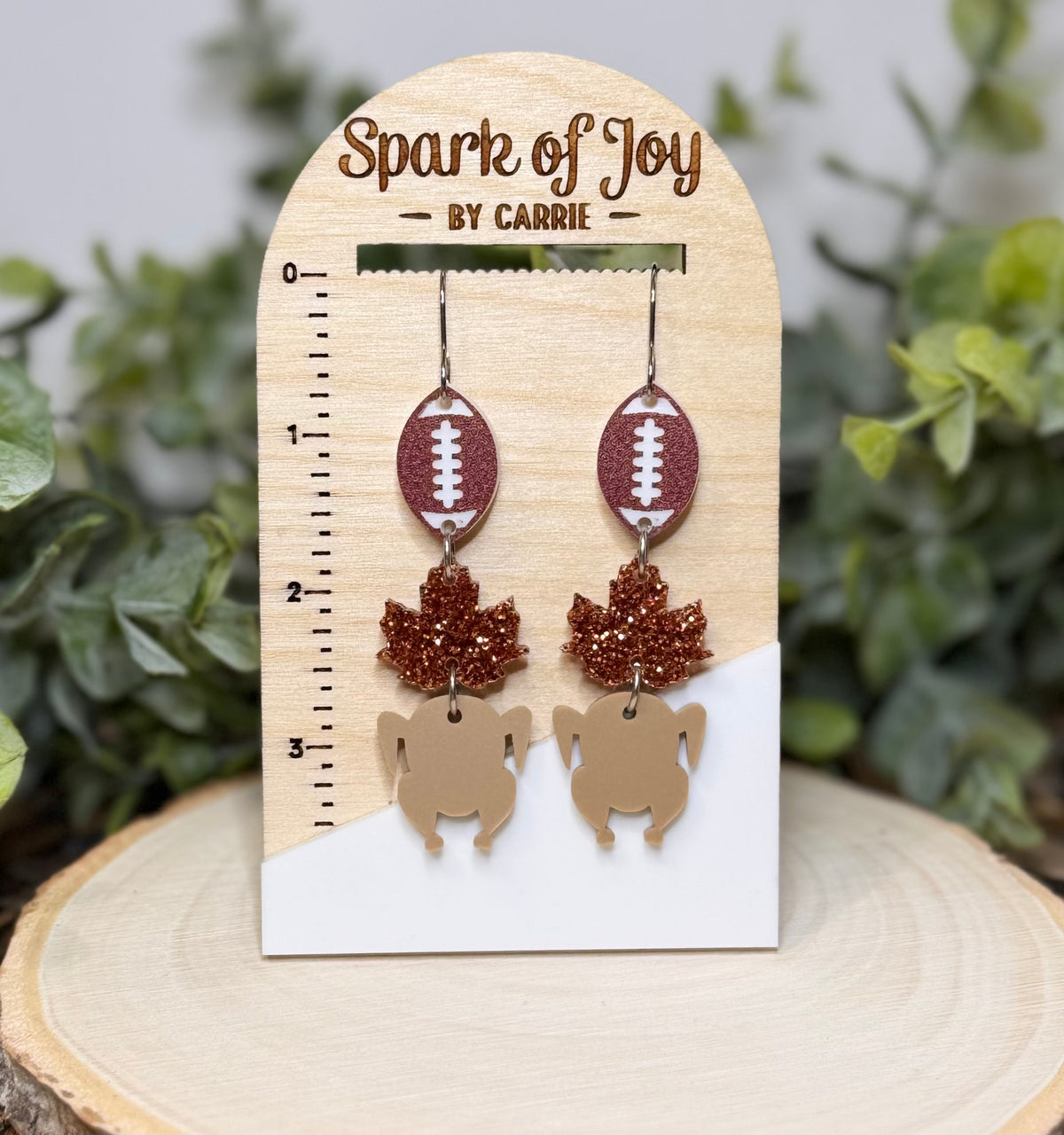 A pair of dangling earrings displayed on a wooden card labeled 'Spark of Joy BY CARrie.' The earrings have football-shaped top pieces in brown glitter with white laces, copper glitter leaf drops, and tan turkey-shaped pendants.