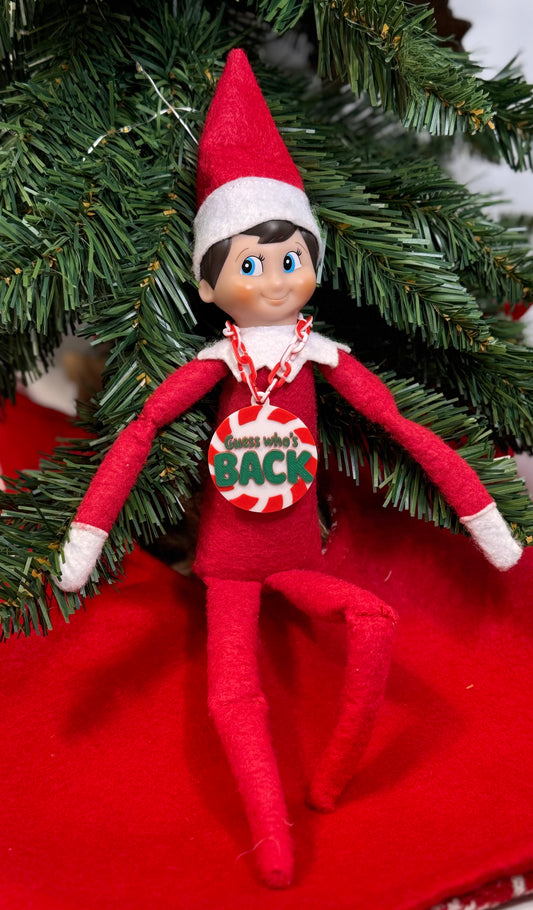 Elf doll wearing a red and white candy cane chain with a peppermint “Guess who’s back” pendant.