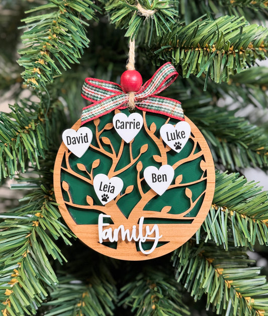 A personalized wooden family tree ornament hanging on a green Christmas tree branch. The circular ornament features a laser-cut tree design with five white heart-shaped nameplates printed with family names (David, Carrie, Luke, Leia, Ben) and a red beaded hanging loop with a red, green, and white plaid ribbon bow.