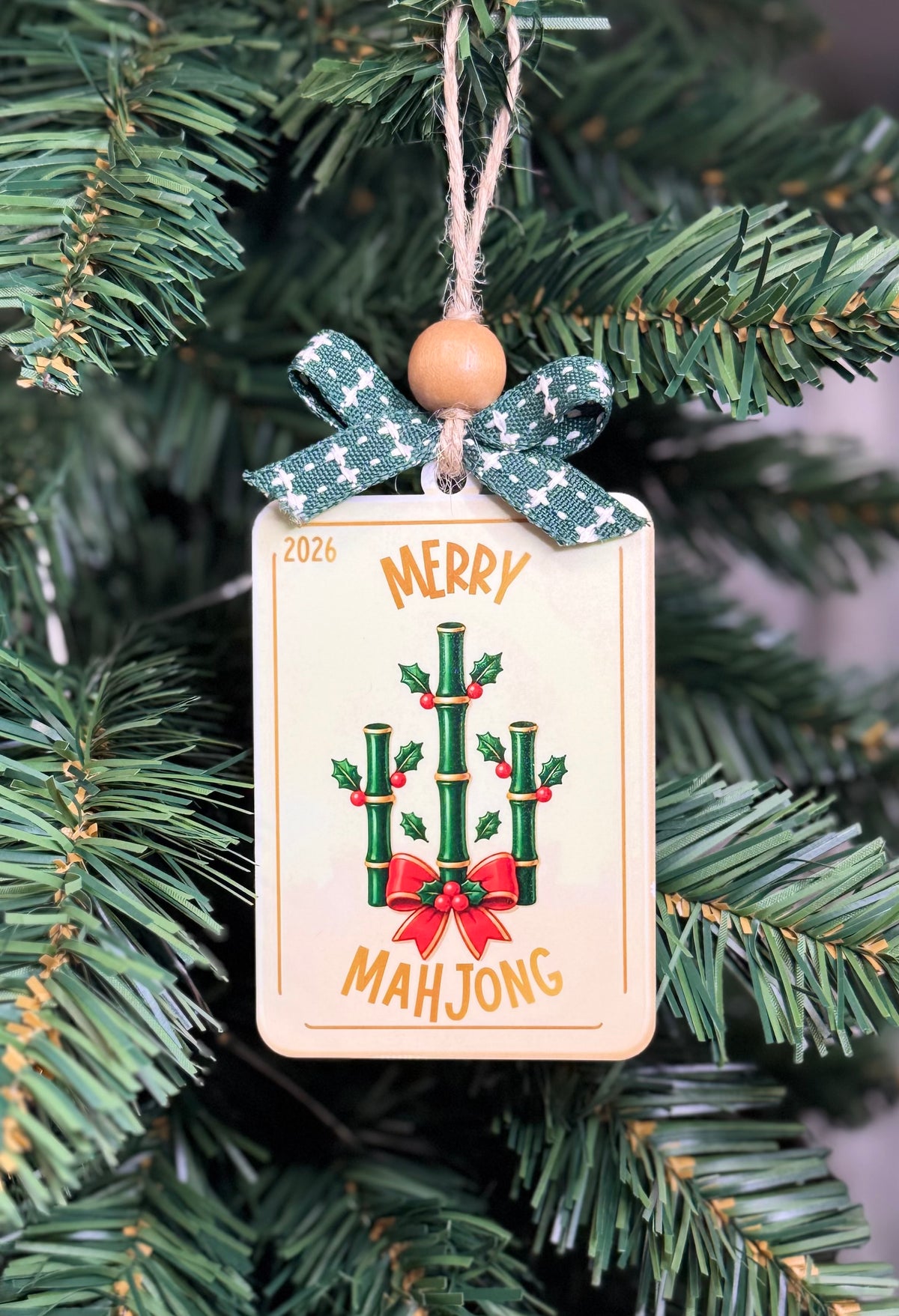 A rectangular Christmas ornament hanging from a green tree branch. The ornament is cream-colored with a printed design of green bamboo stalks, red bows, and the words 'MERRY 2026 MAHJONG' in gold.