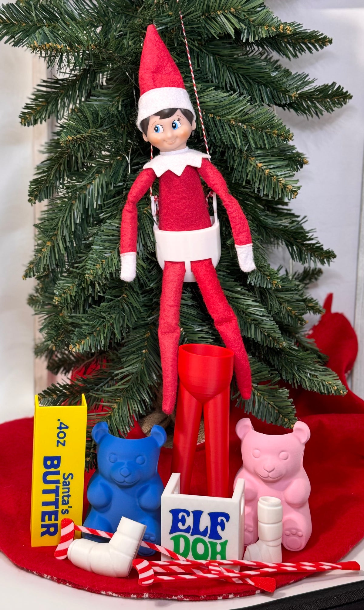 Elf doll sitting in a white 3D printed swing hung by red and white twine in a Christmas tree.
