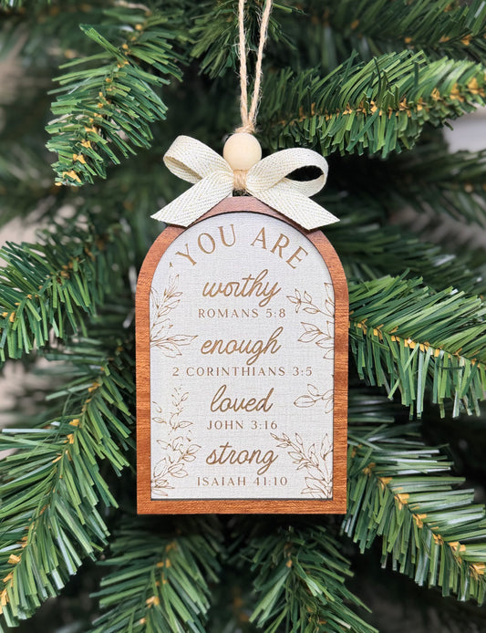 A wooden Christmas ornament shaped like a book cover hangs from a tree branch. The ornament has a cream ribbon bow at the top, a light wood bead, and a wood-grain border.