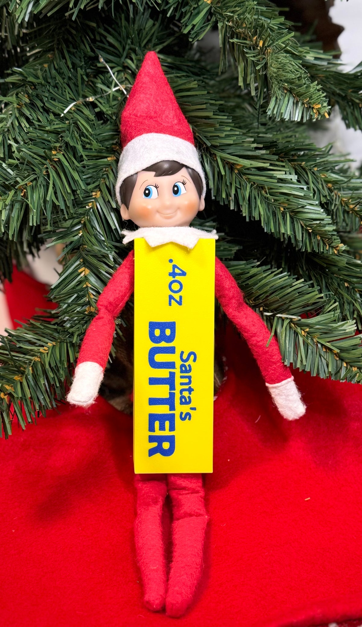 Elf doll wearing a yellow 3D printed butter stick costume with blue “Santa’s Butter” lettering, sitting under a Christmas tree