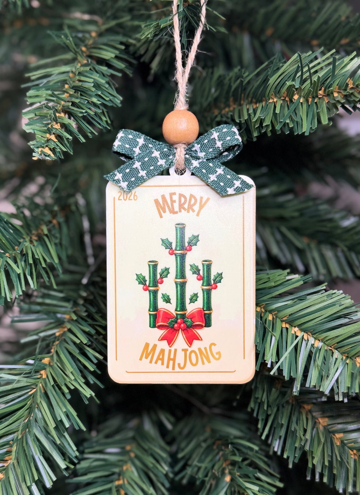 A rectangular Christmas ornament hanging from a green tree branch. The ornament has a cream background with a bamboo and red bow graphic, the words “MERRY MAHJONG,” and a green patterned ribbon tied around the top with a wooden bead.
