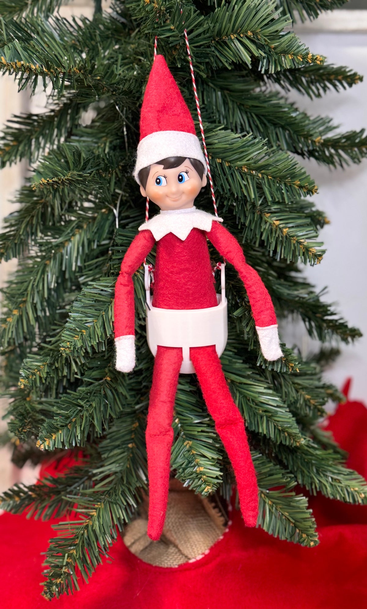 Elf doll sitting in a white 3D printed swing hung by red and white twine in a Christmas tree.