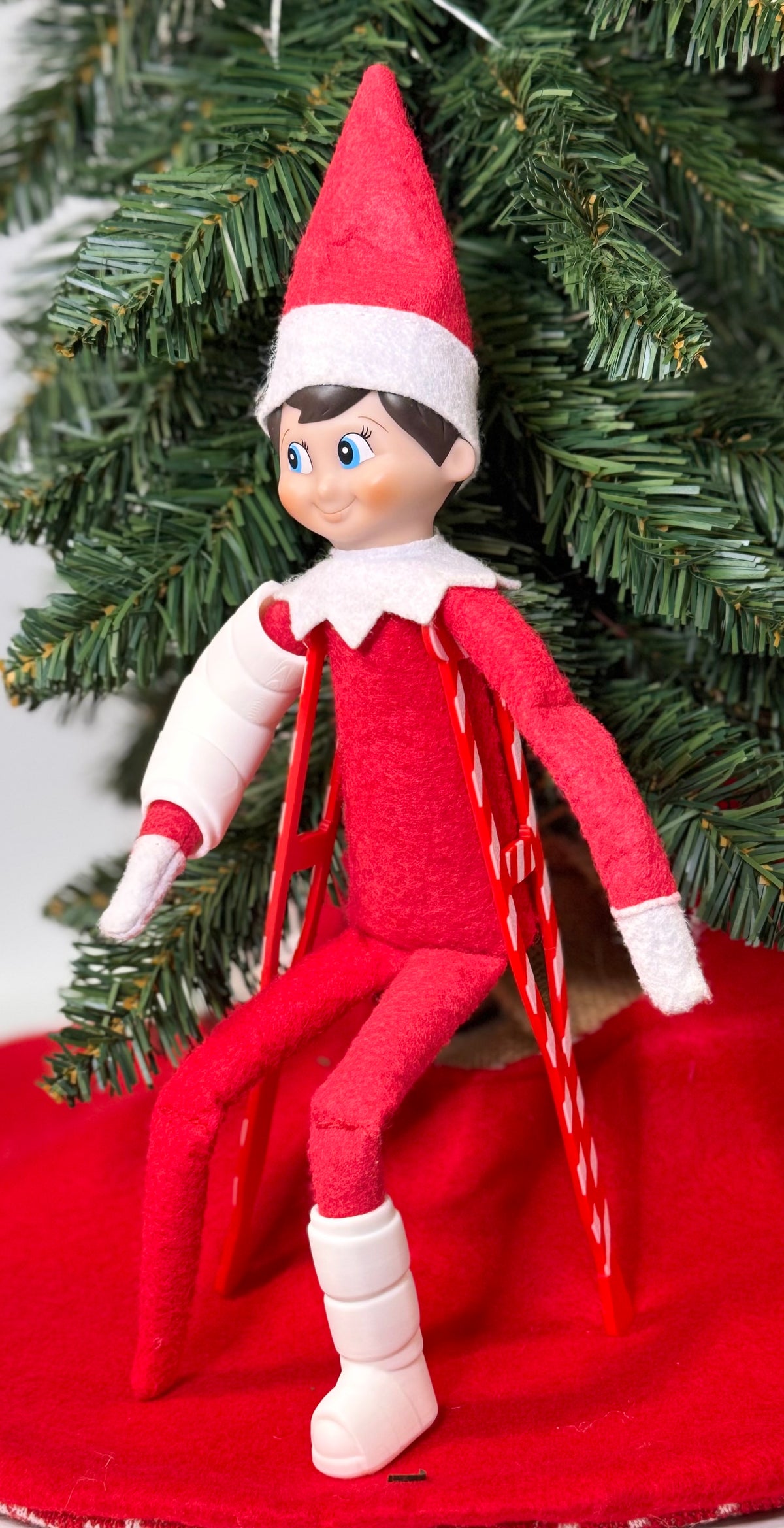 Elf doll wearing a white 3D printed arm cast and leg cast, standing with red crutches under a Christmas tree