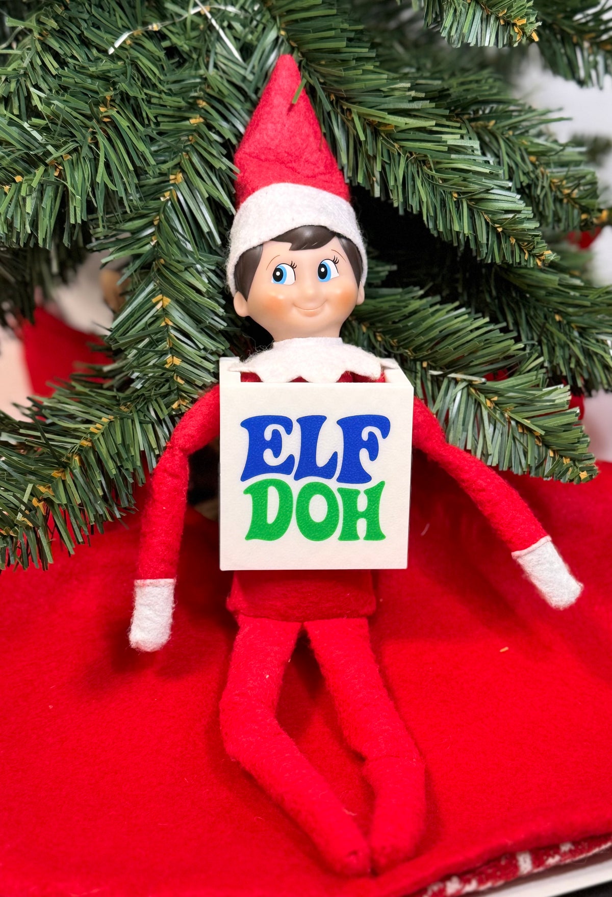Elf doll wearing a white 3D printed box costume with blue and green “Elf Doh” lettering, sitting under a Christmas tree.