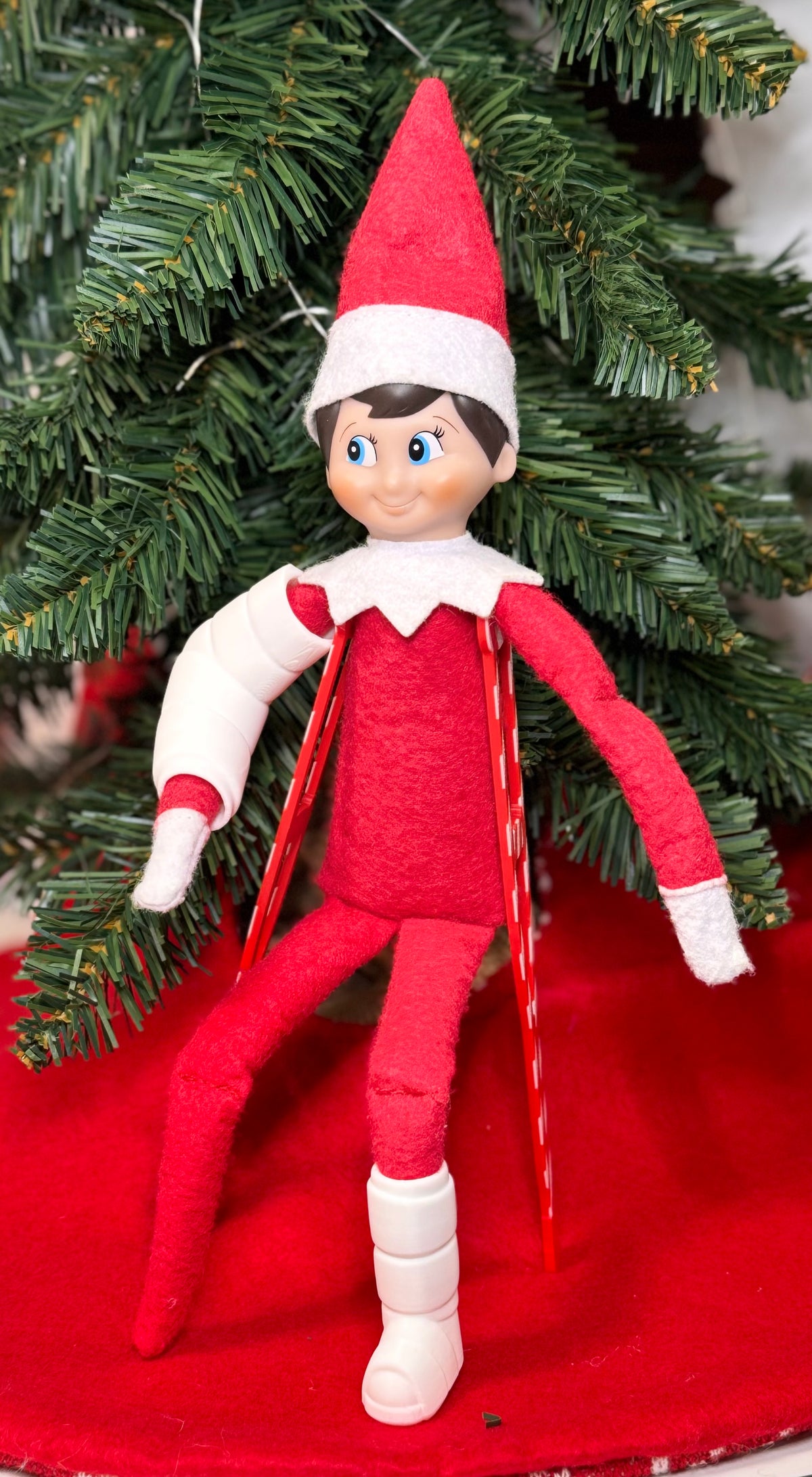 Elf doll wearing a white 3D printed arm cast and leg cast, standing with red crutches under a Christmas tree