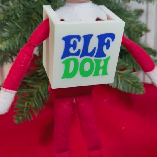Elf doll wearing a white 3D printed box costume with blue and green “Elf Doh” lettering, sitting under a Christmas tree.