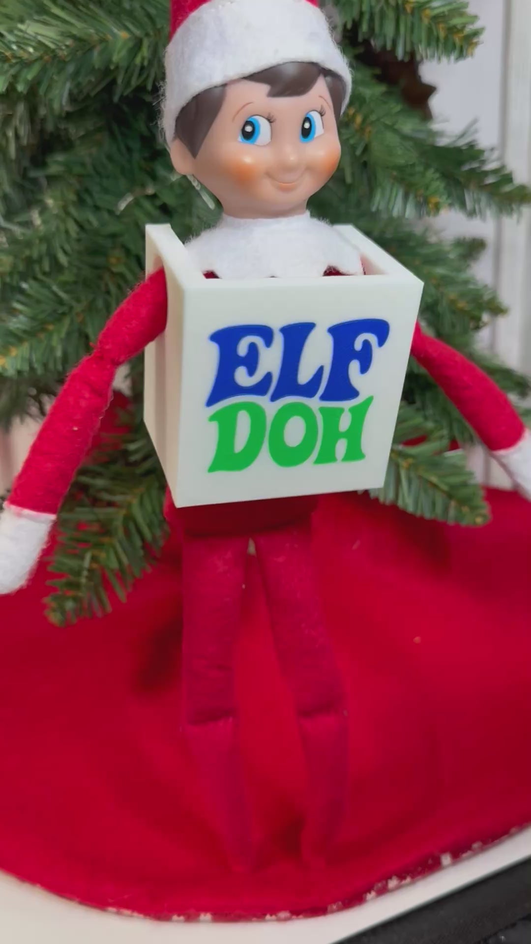 Elf doll wearing a white 3D printed box costume with blue and green “Elf Doh” lettering, sitting under a Christmas tree.