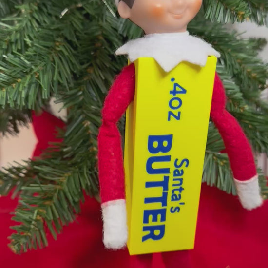 Elf doll wearing a yellow 3D printed butter stick costume with blue “Santa’s Butter” lettering, sitting under a Christmas tree
